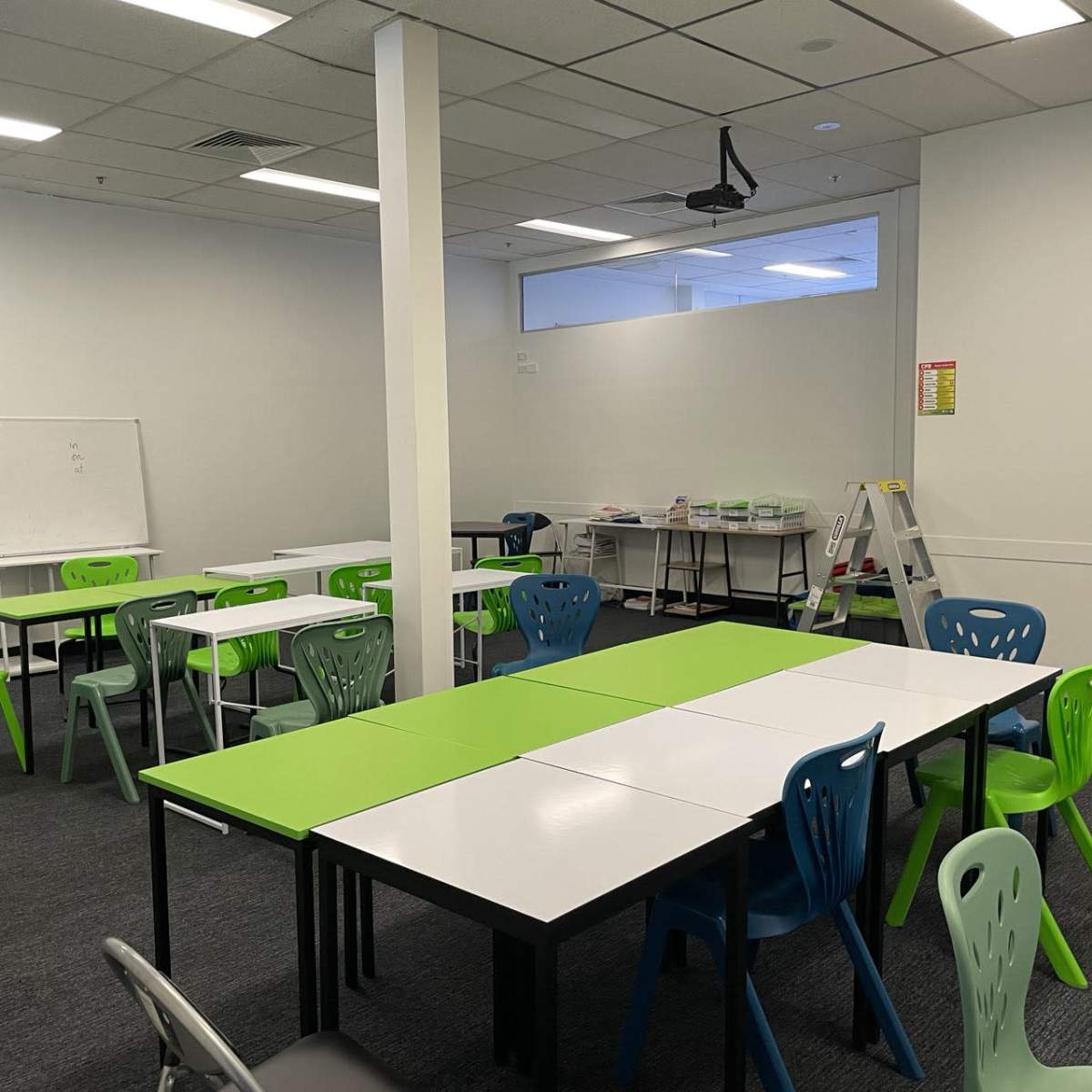 A classroom setting features multiple tables with green and white surfaces, accompanied by colorful chairs and a whiteboard, all arranged in a spacious area.