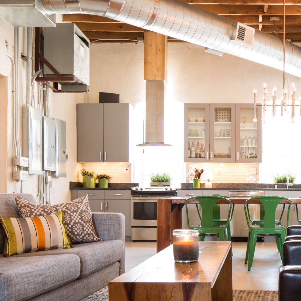 A modern kitchen and living area features stainless steel appliances, green metal chairs, and wooden furniture under exposed beams and ductwork.