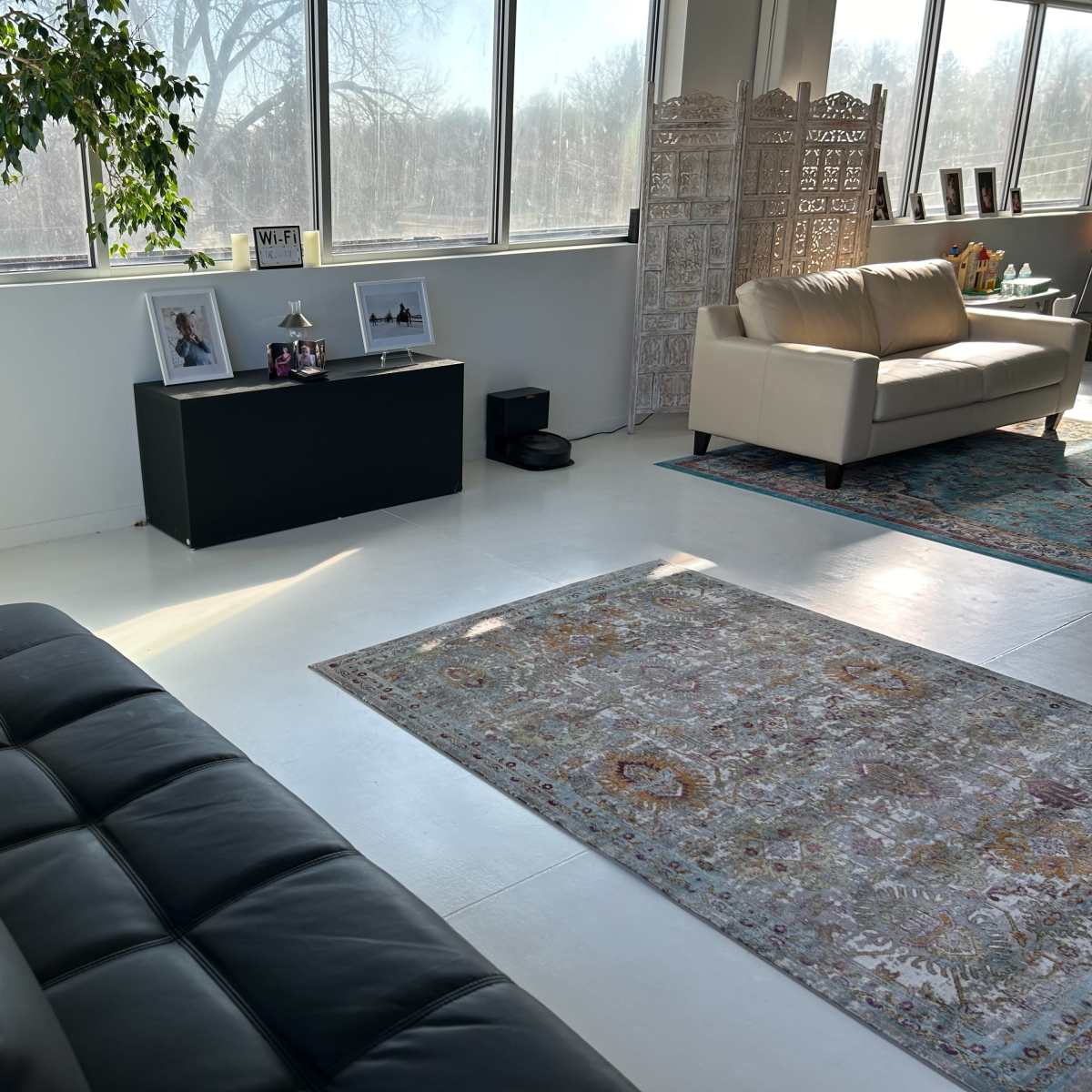 A spacious, bright living room with large windows, a black sofa, a decorative area rug, and minimal furnishings including a black entertainment unit and a cream-colored couch.