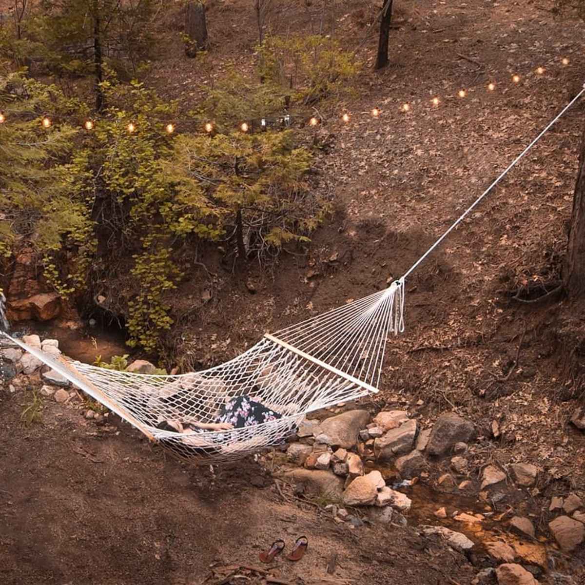 A white hammock is suspended between two trees above a rocky creek in a wooded area, with string lights hanging overhead.