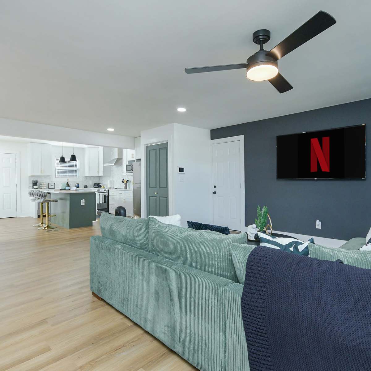 A spacious living room features a green sectional sofa, a ceiling fan, and a large television displaying the Netflix logo.