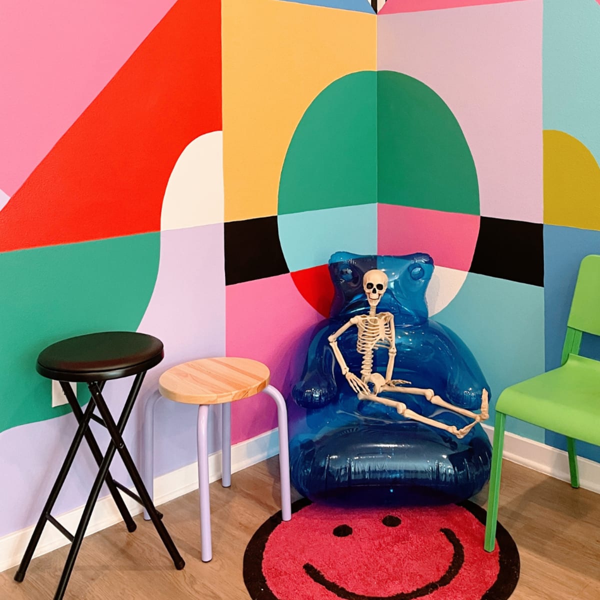 A skeleton sits in a blue inflatable chair in a colorful corner decorated with abstract shapes and vibrant colors, surrounded by three different chairs and a smiley face rug.