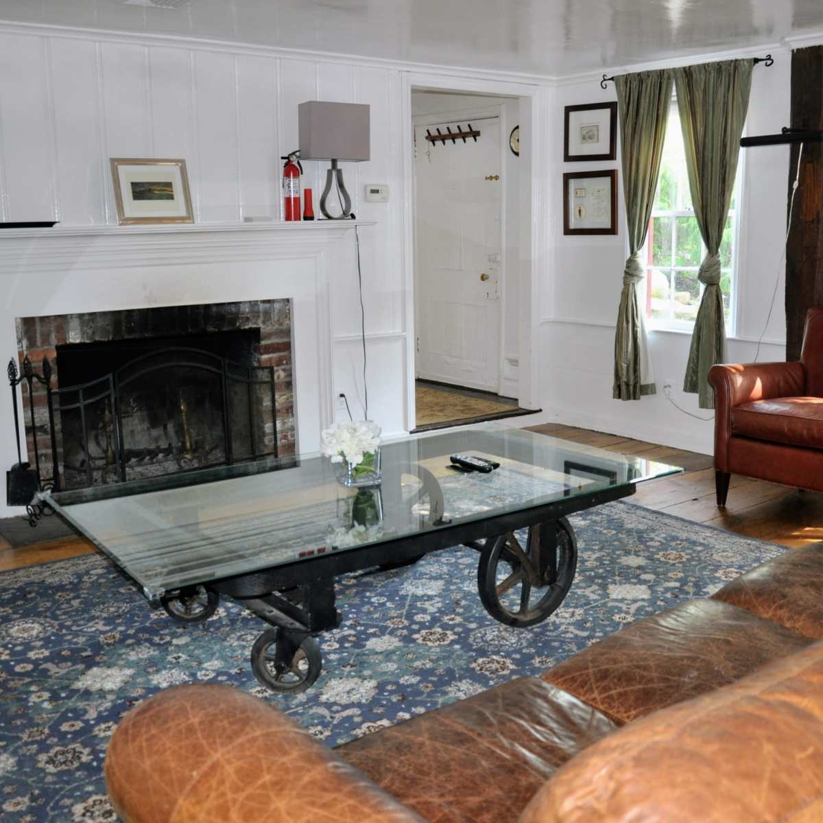The living room features a glass coffee table on wheels, a brown leather sofa, an armchair, and a fireplace flanked by windows with curtains.