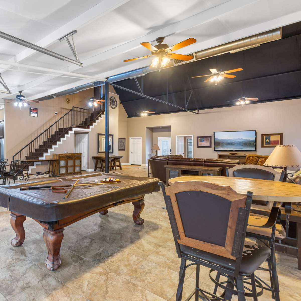 A large, open space featuring a billiard table, dining tables, and a staircase in a well-lit, multi-purpose room.