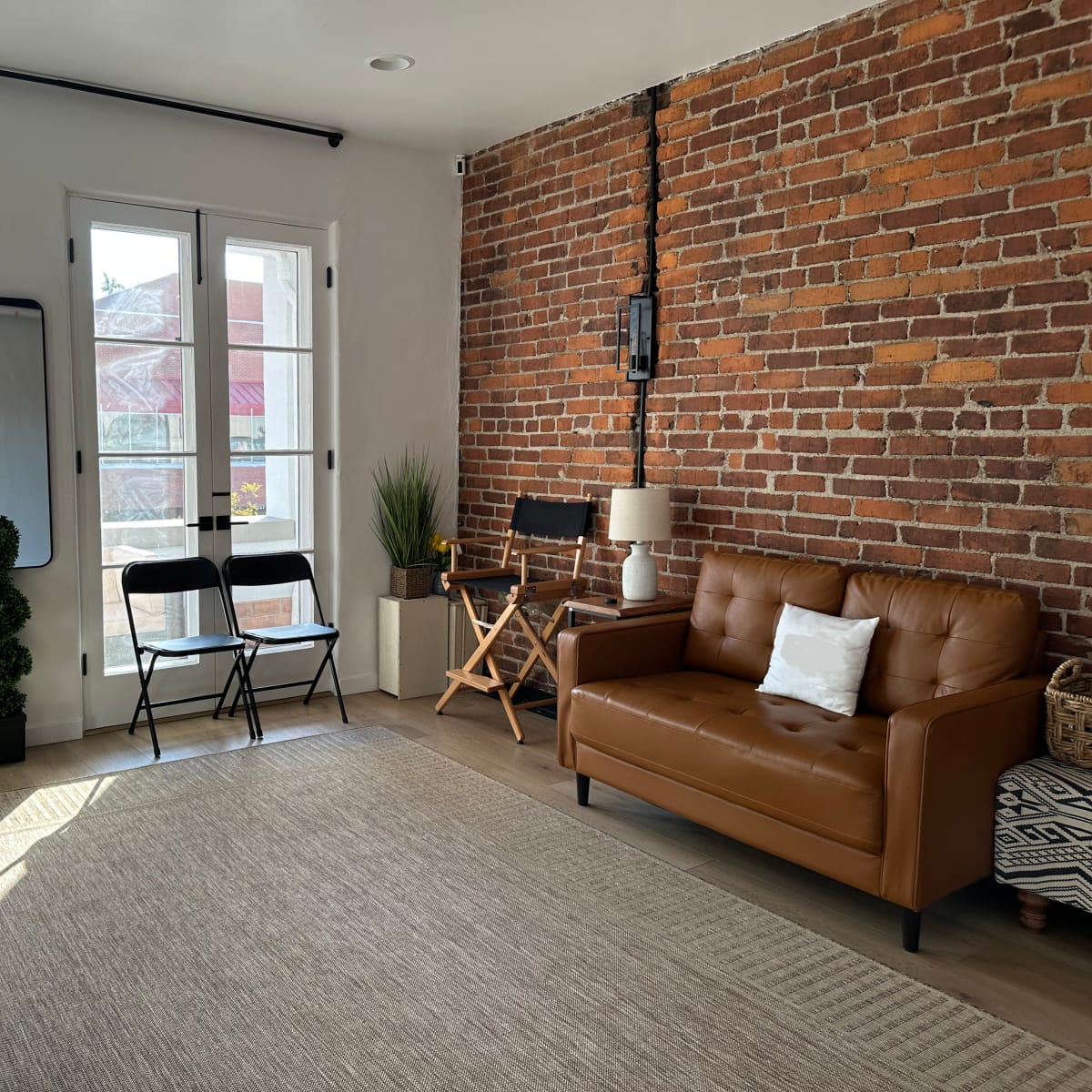 A room with exposed brick walls, featuring a leather couch, a chair, a potted plant, and several black folding chairs arranged along the wall beside large windows.