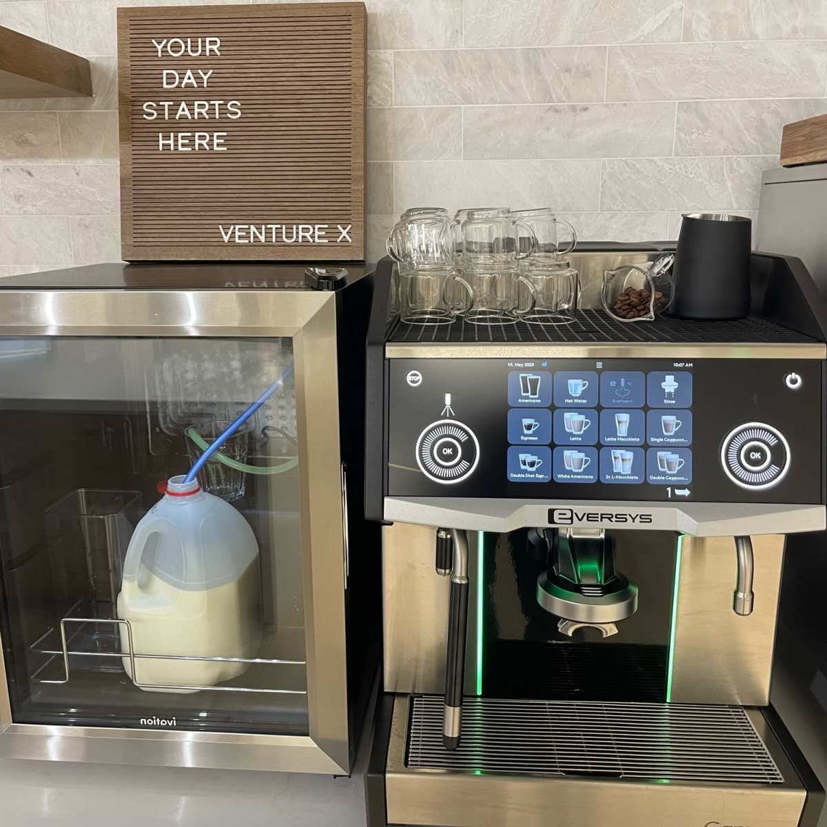 A milk dispenser and a coffee machine are placed side by side on a counter with a sign above that reads "YOUR DAY STARTS HERE" and "VENTURE X."