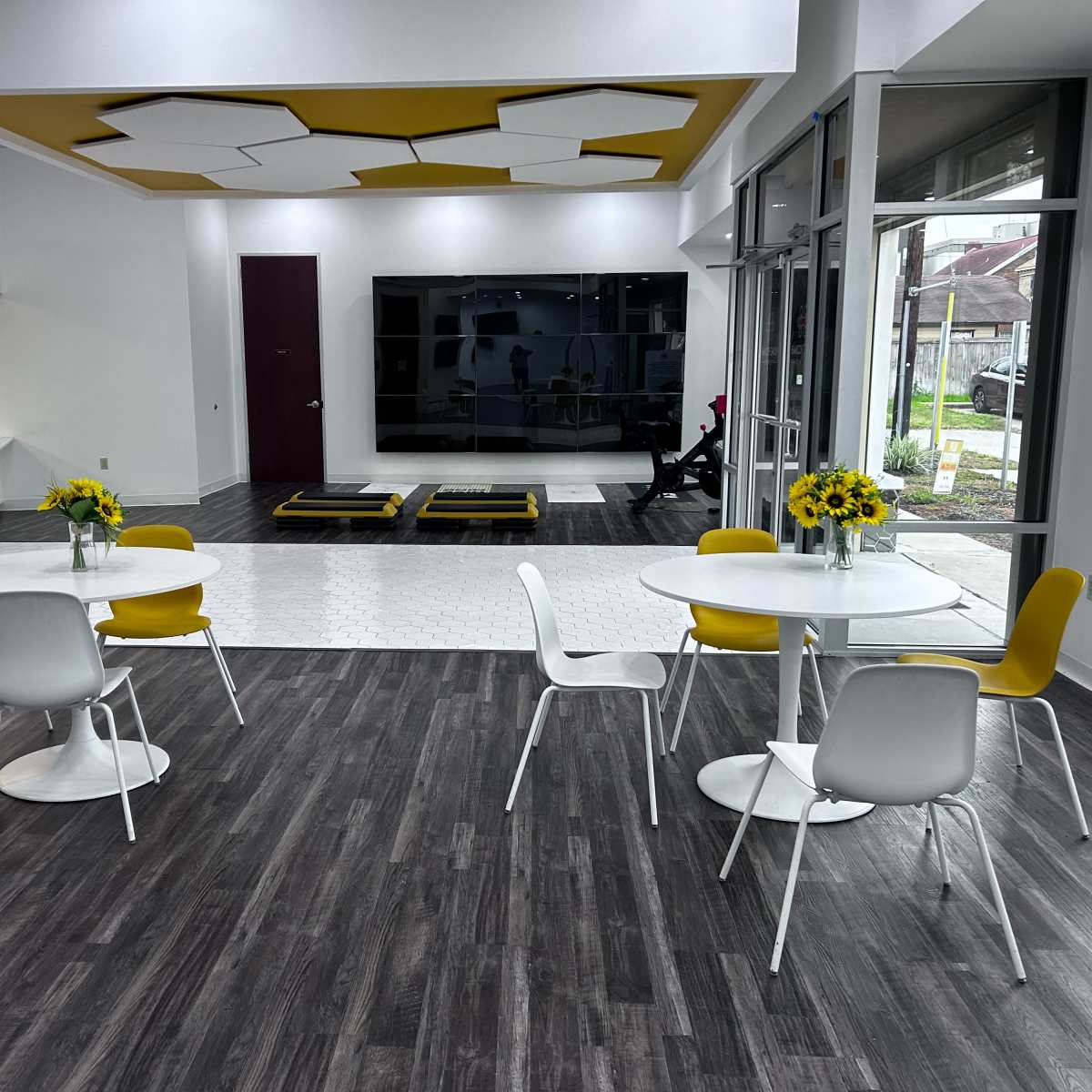 A modern, open space featuring two round tables with yellow chairs, a large wall-mounted screen, and a section with a patterned white floor.