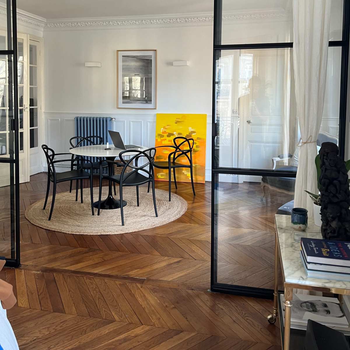 A modern interior space with a round table and black chairs, a yellow abstract painting on the wall, and a glass partition separating the room.