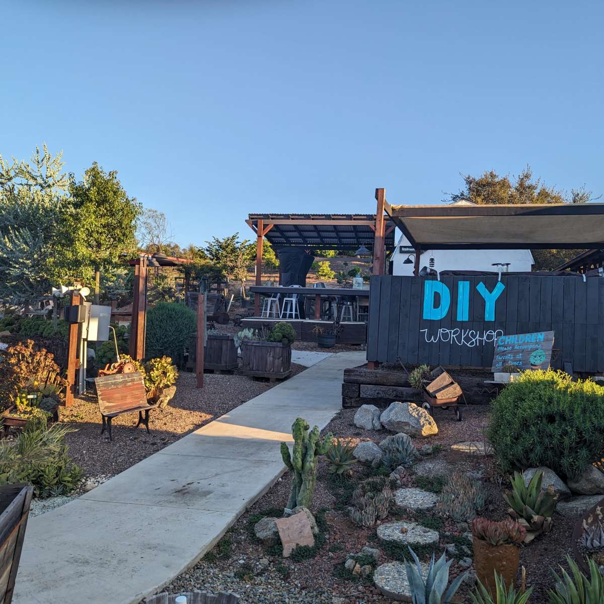 A DIY workshop area with a path leading to a structure surrounded by various plants and outdoor furniture.