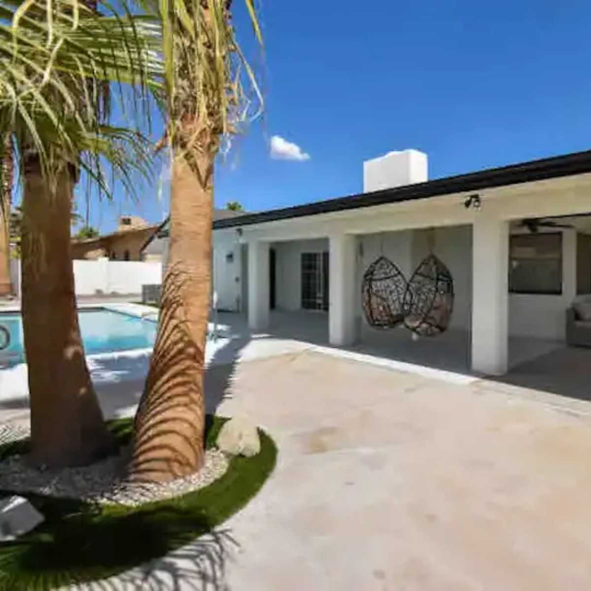 A modern backyard with a swimming pool, palm trees, and a covered seating area featuring a hanging chair.