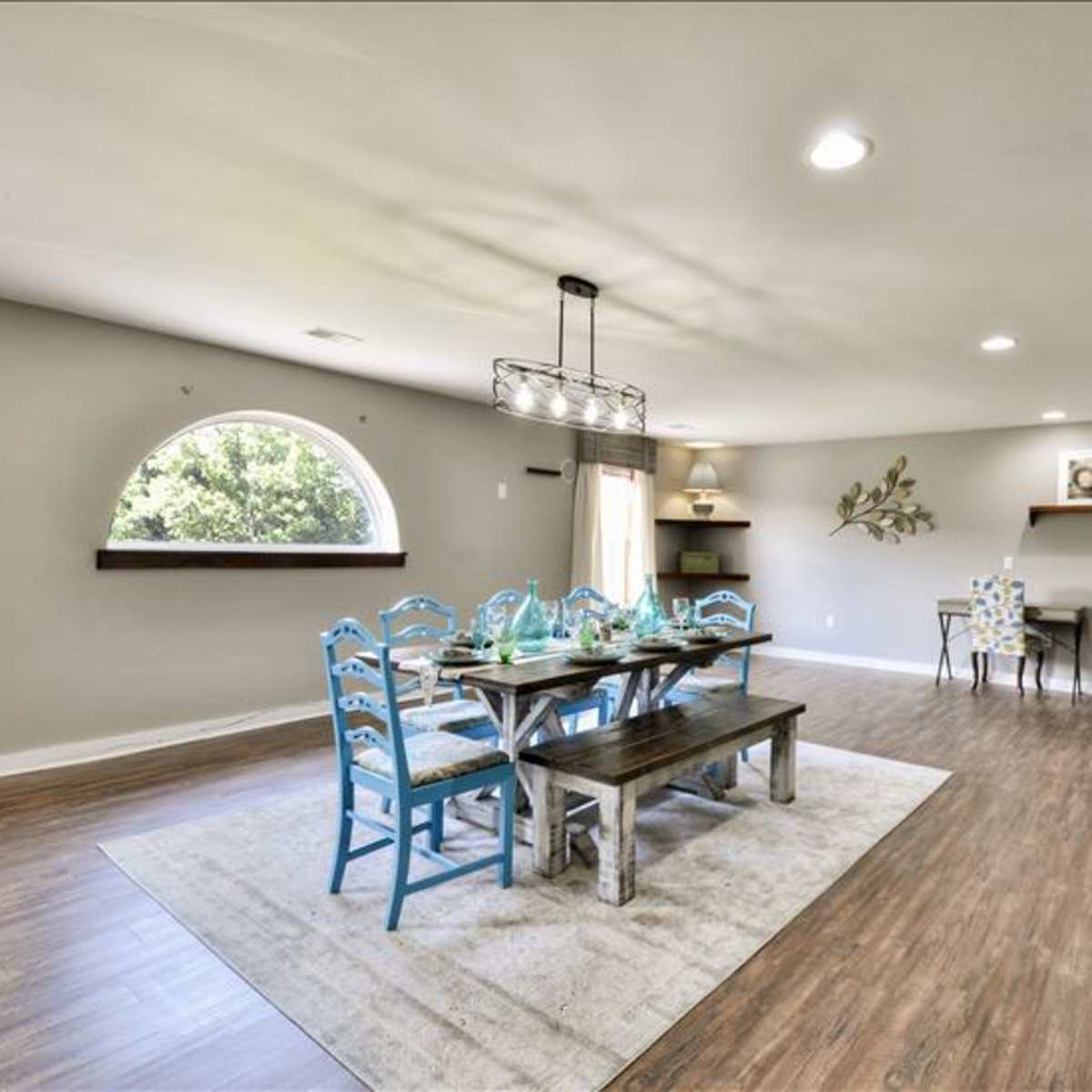 A spacious dining area featuring a long wooden table with blue chairs, an oval window, and minimalist decor against light-colored walls.