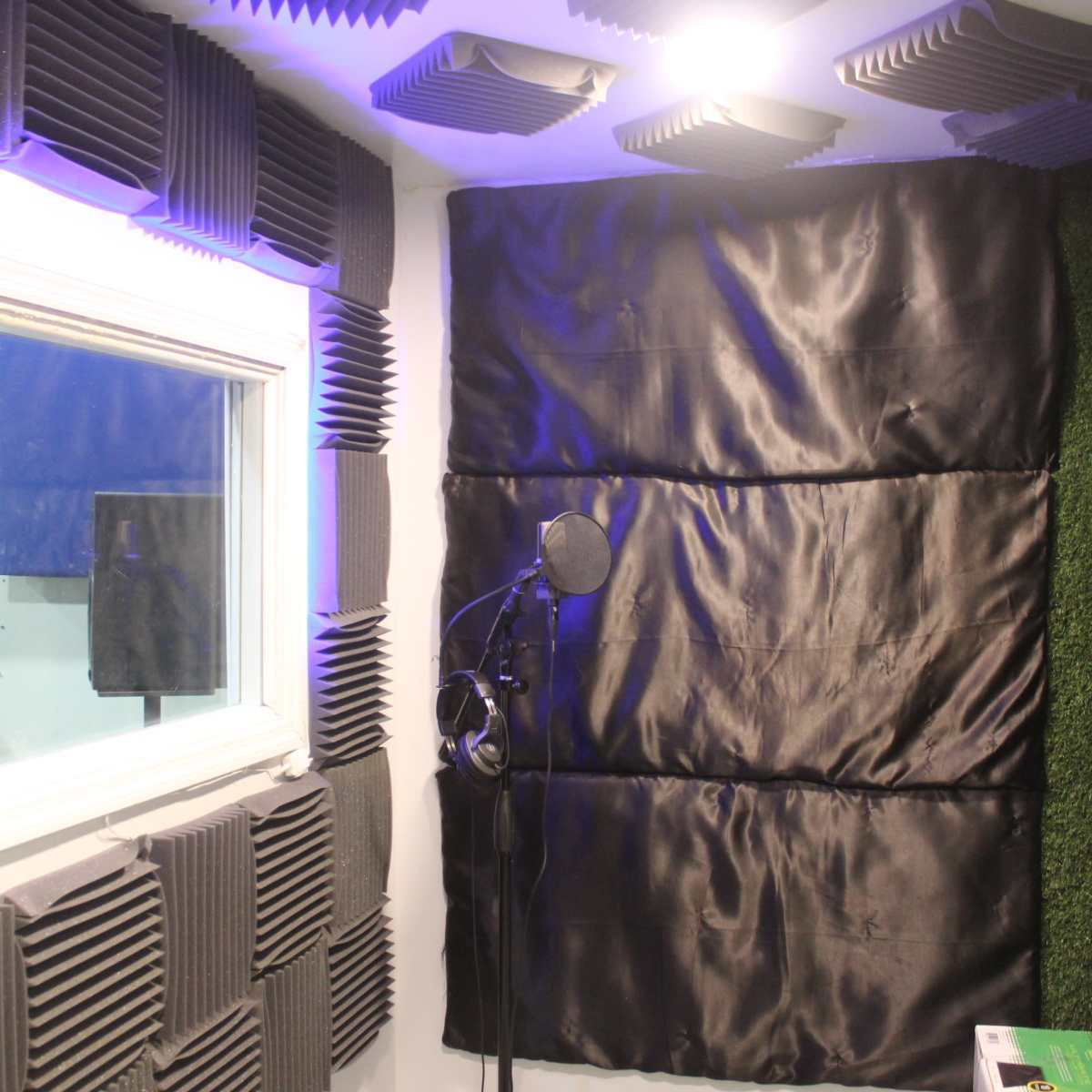 A soundproof recording booth with acoustic foam panels, a microphone on a stand, and a window for visual access.