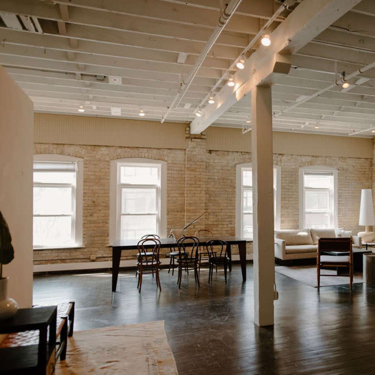 An open-concept living space with exposed brick walls, large windows, and distinct areas for a dining table, a lounge, and a sitting area.