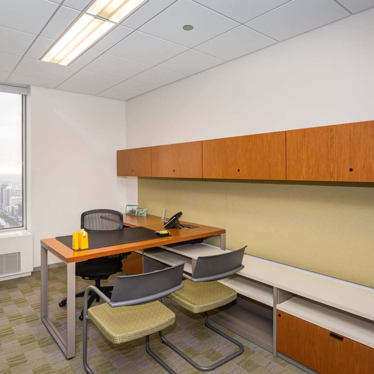 A modern office space with a desk, two chairs, and storage cabinets, featuring a large window overlooking a city skyline.