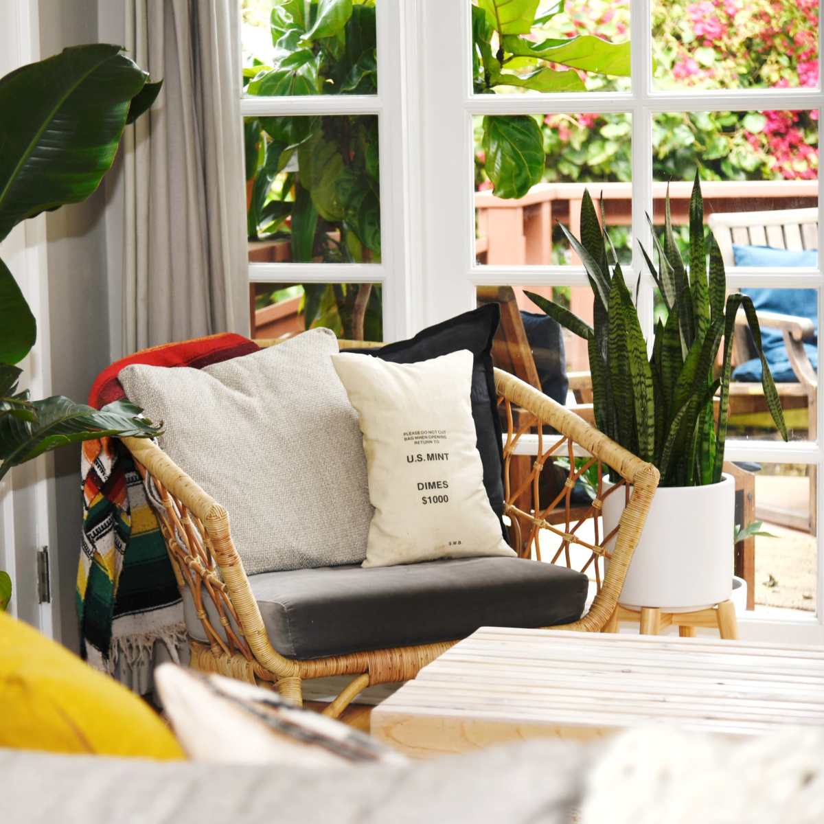A cozy seating area features a rattan chair with cushions, surrounded by potted plants and large windows overlooking an outdoor space.