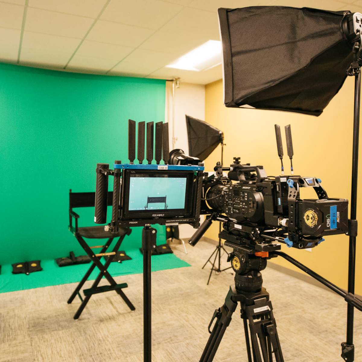 A video production setup featuring a camera on a tripod, a green backdrop, and studio lighting equipment positioned around a director's chair.