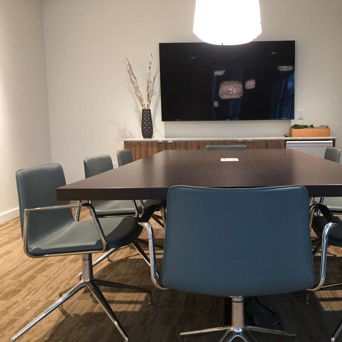 A modern conference room features a large rectangular table surrounded by several chairs and a wall-mounted screen.