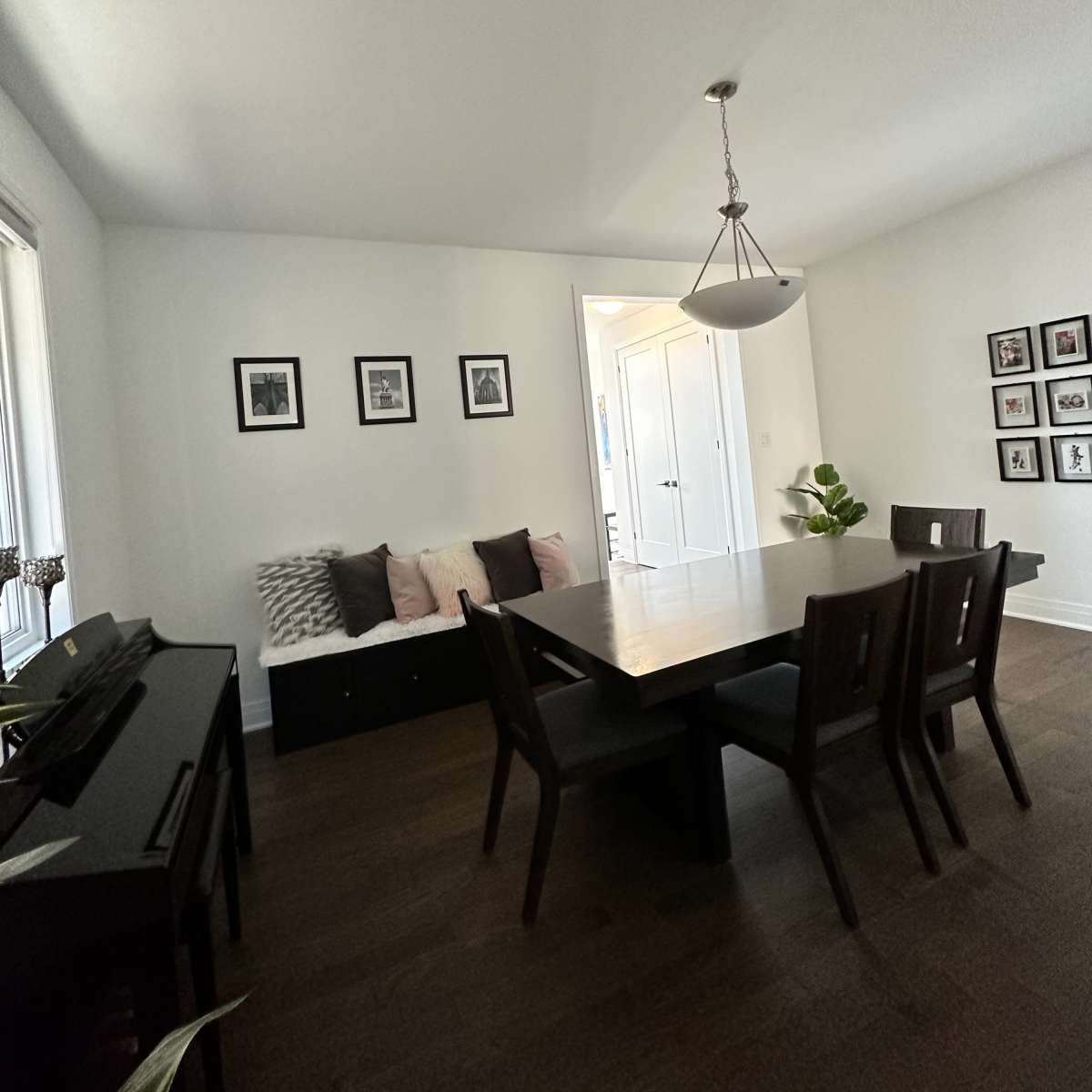 A dining area with a rectangular table surrounded by dark wooden chairs, a black bench with cushions against the wall, and framed photos hung on the light-colored walls.