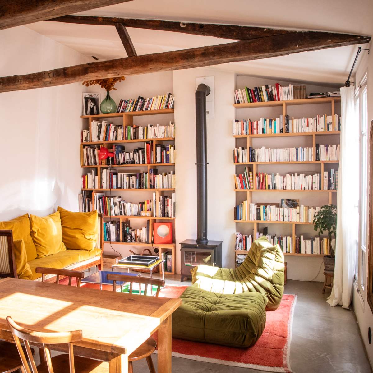 A cozy living room features a wooden table and chairs, a green beanbag chair, shelves filled with books, and sunlight streaming through a window.