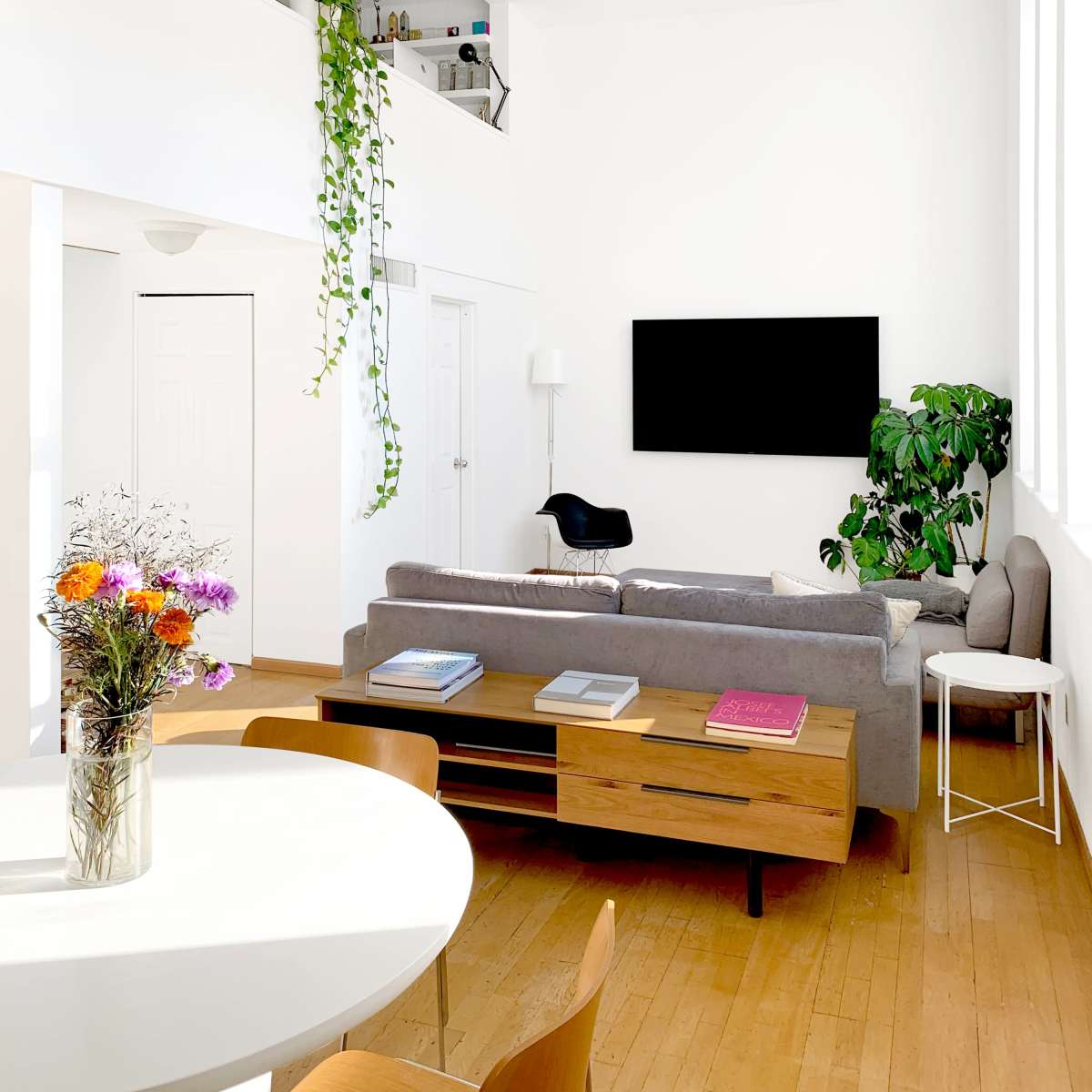 A bright living room featuring a gray sofa, a wooden coffee table, a wall-mounted TV, and large windows that allow natural light to enter the space.