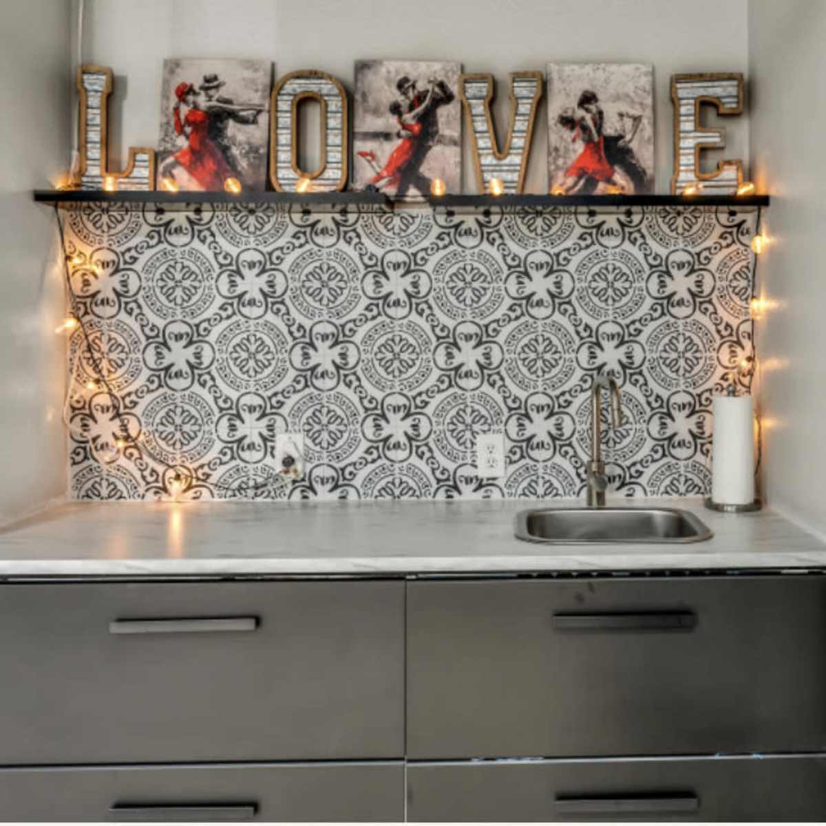 A small kitchen area with a sink, dark cabinets, decorative letters spelling "LOVE," and a patterned backsplash illuminated by string lights.