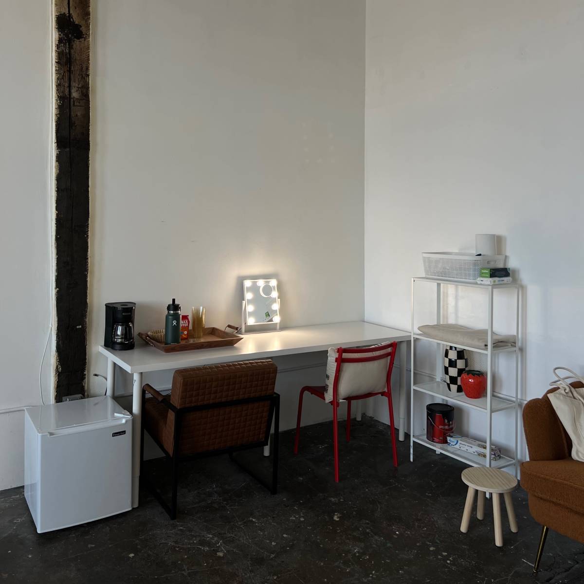 A minimal workspace features a white table with two chairs, a coffee maker, and shelves, set against a stark white wall.