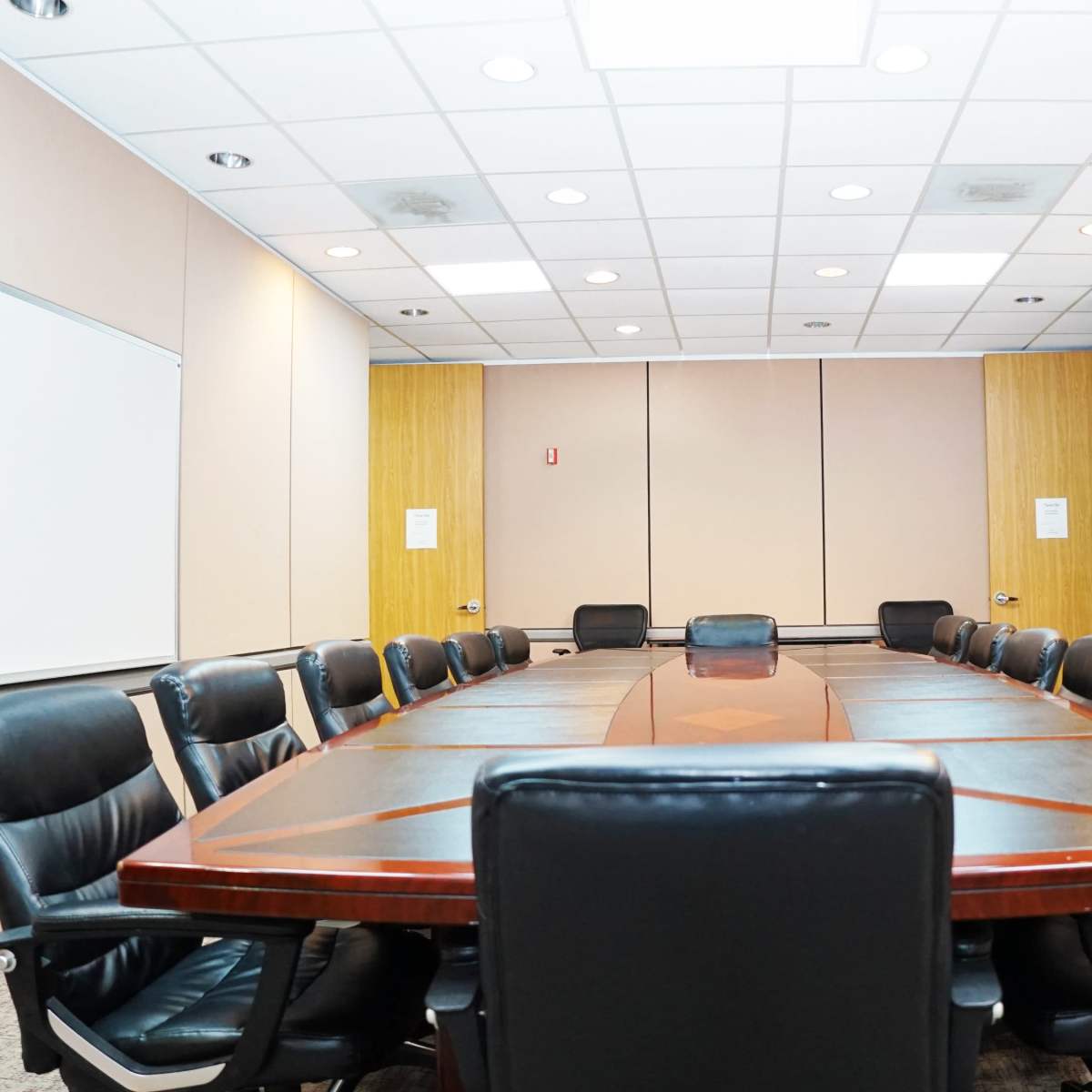 A long wooden conference table surrounded by black leather chairs is set in a sparsely decorated meeting room with beige walls and a whiteboard.