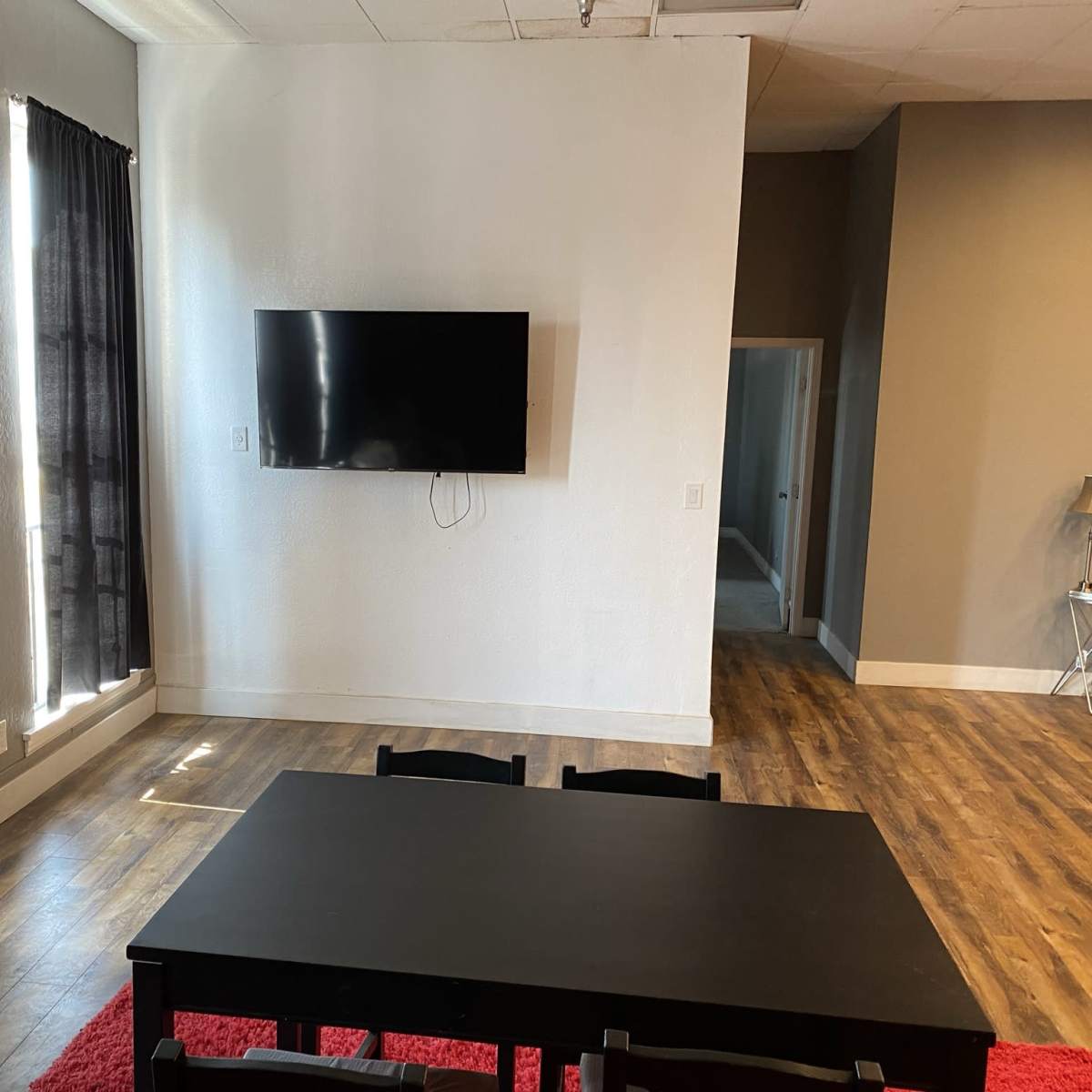A black table is positioned in a well-lit room with a television mounted on the wall and dark curtains covering the windows.