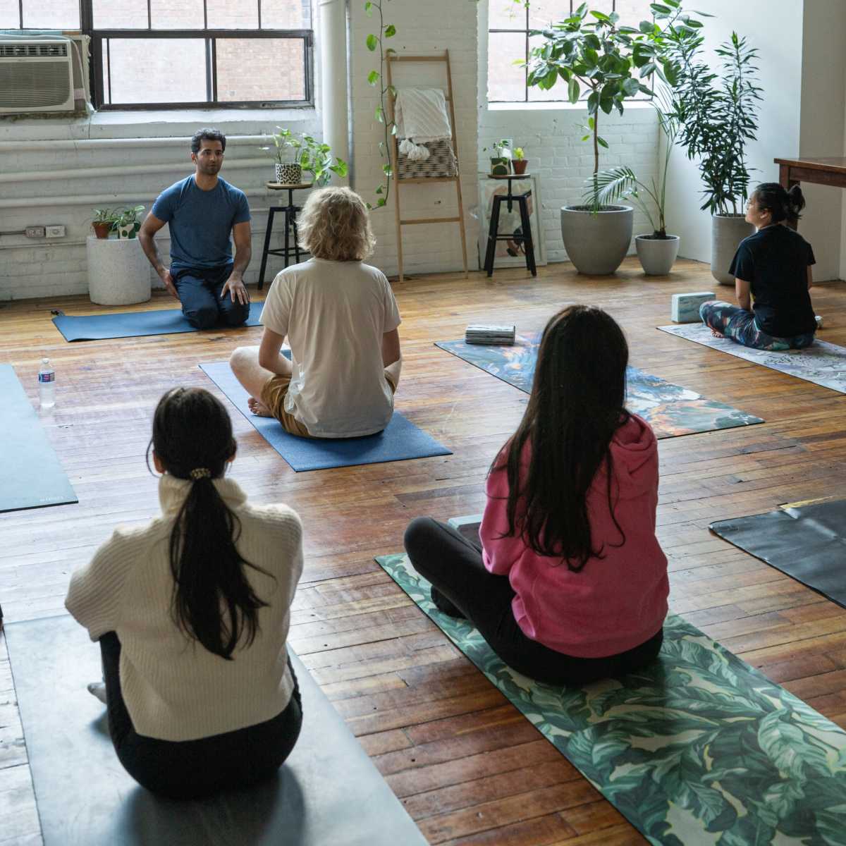 A yoga instructor leads a class in a bright, plant-filled studio with participants seated on mats.