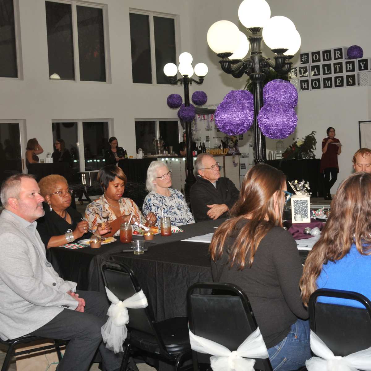 A group of people sits around a large table in a well-lit room decorated with purple accents and a chandelier.