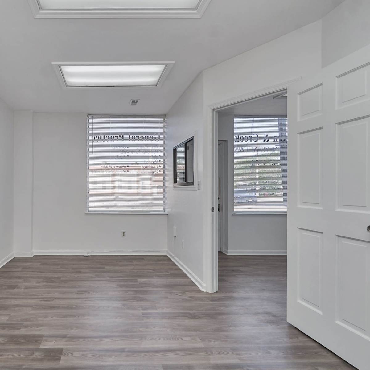 A vacant office space with white walls, hardwood-like flooring, and a door leading to another room.