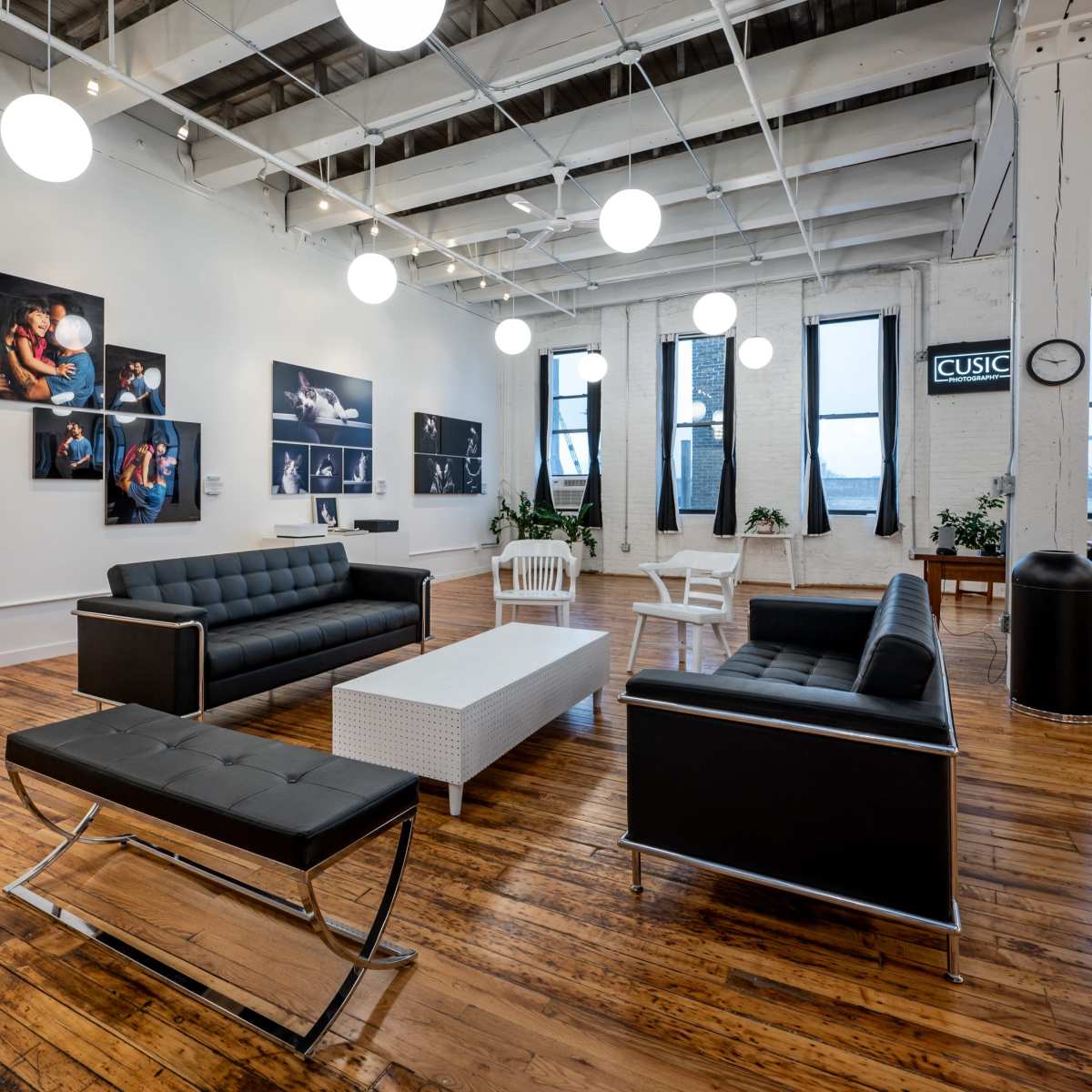 A modern interior space with black leather sofas, a white coffee table, and several framed photographs on the white walls, illuminated by hanging round light fixtures.