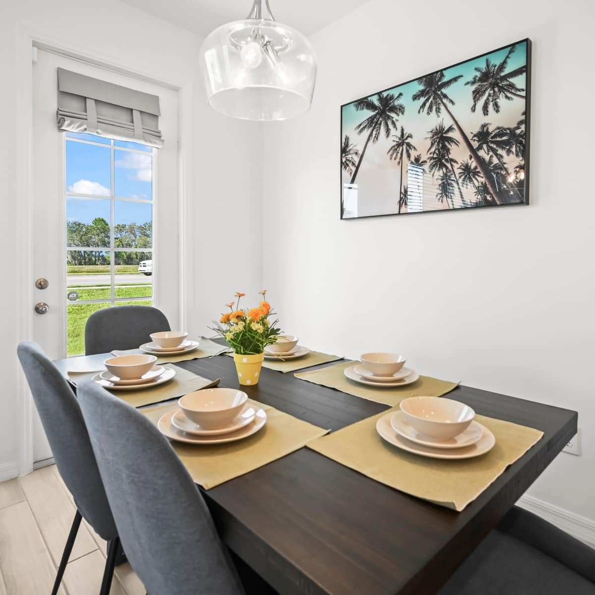 A dining table is set with plates and a flower centerpiece in a bright room featuring a window with a view of greenery.