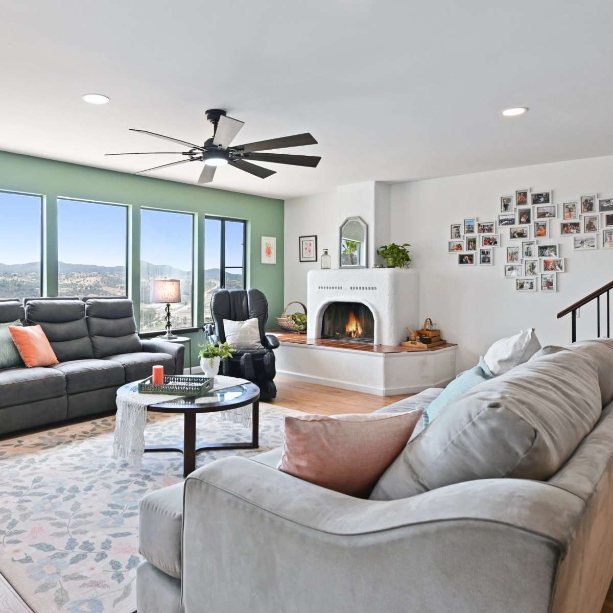 The living room features two sofas, a circular coffee table, and large windows with a view, alongside a fireplace and a wall adorned with photographs.