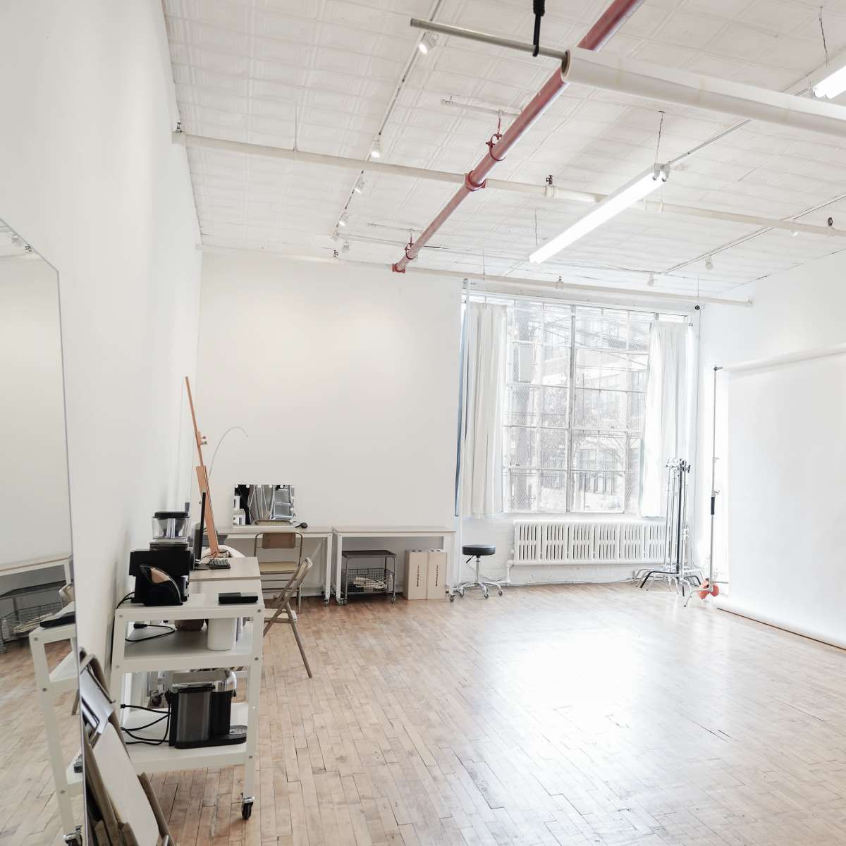 A spacious room with wooden flooring, large windows, a white backdrop on a stand, and various furniture pieces along the walls.