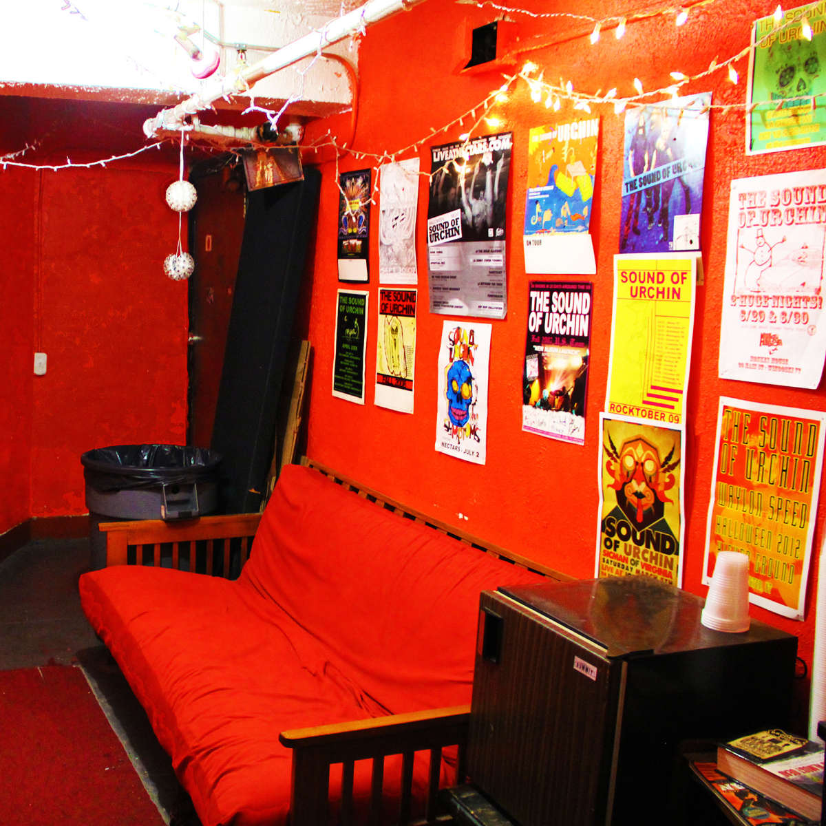 A small, orange-walled room features a red couch, a trash can, and numerous posters on the walls.