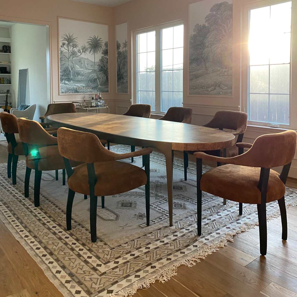 A long wooden dining table surrounded by brown upholstered chairs is set in a brightly lit room with large windows and mural-covered walls.
