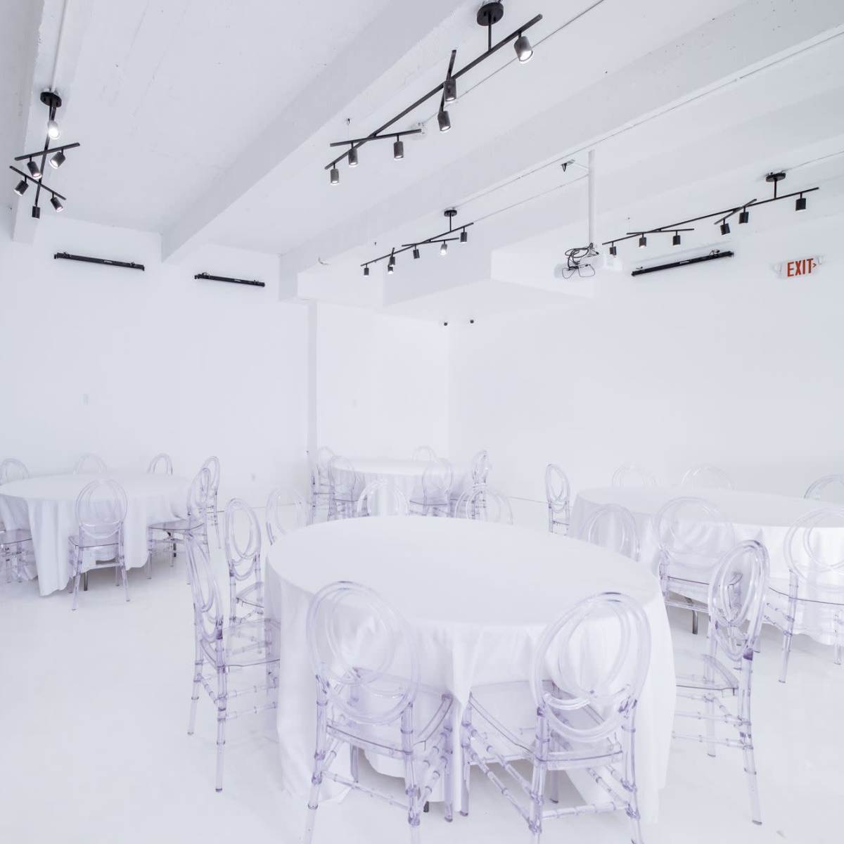 A spacious, white event room with several circular tables covered in white tablecloths and surrounded by clear plastic chairs.