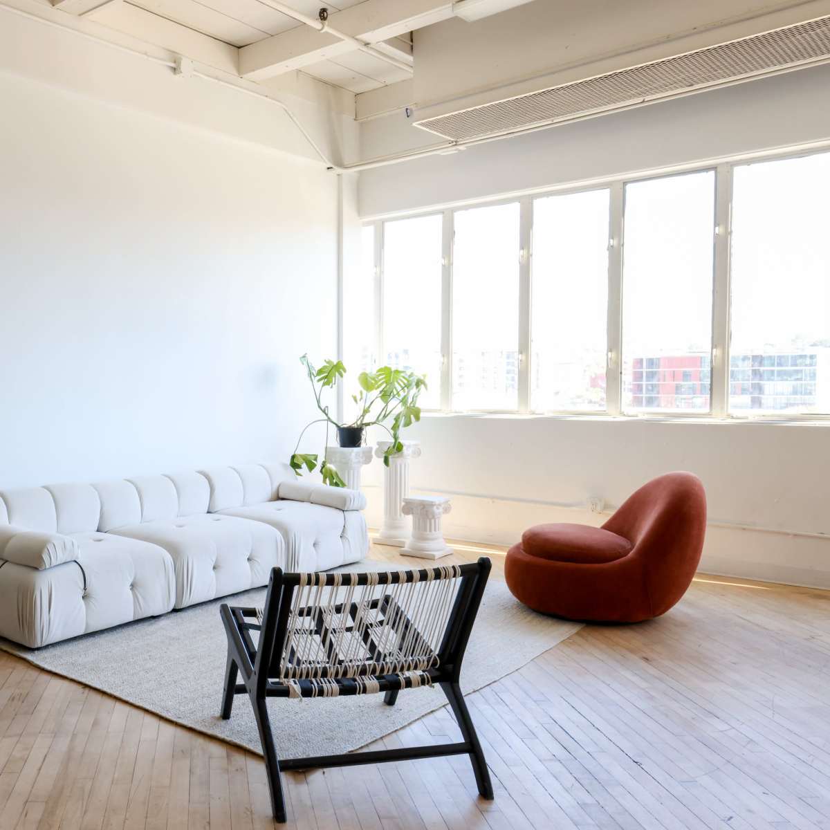 A spacious room with hardwood floors, featuring a beige sofa, a red accent chair, a black woven chair, a white side table, and a large mirror reflecting the sunlight from the windows.