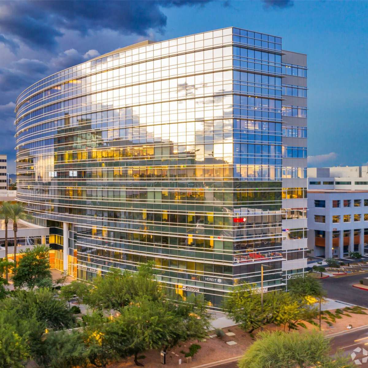 A modern glass office building surrounded by palm trees and additional structures under a cloudy sky.