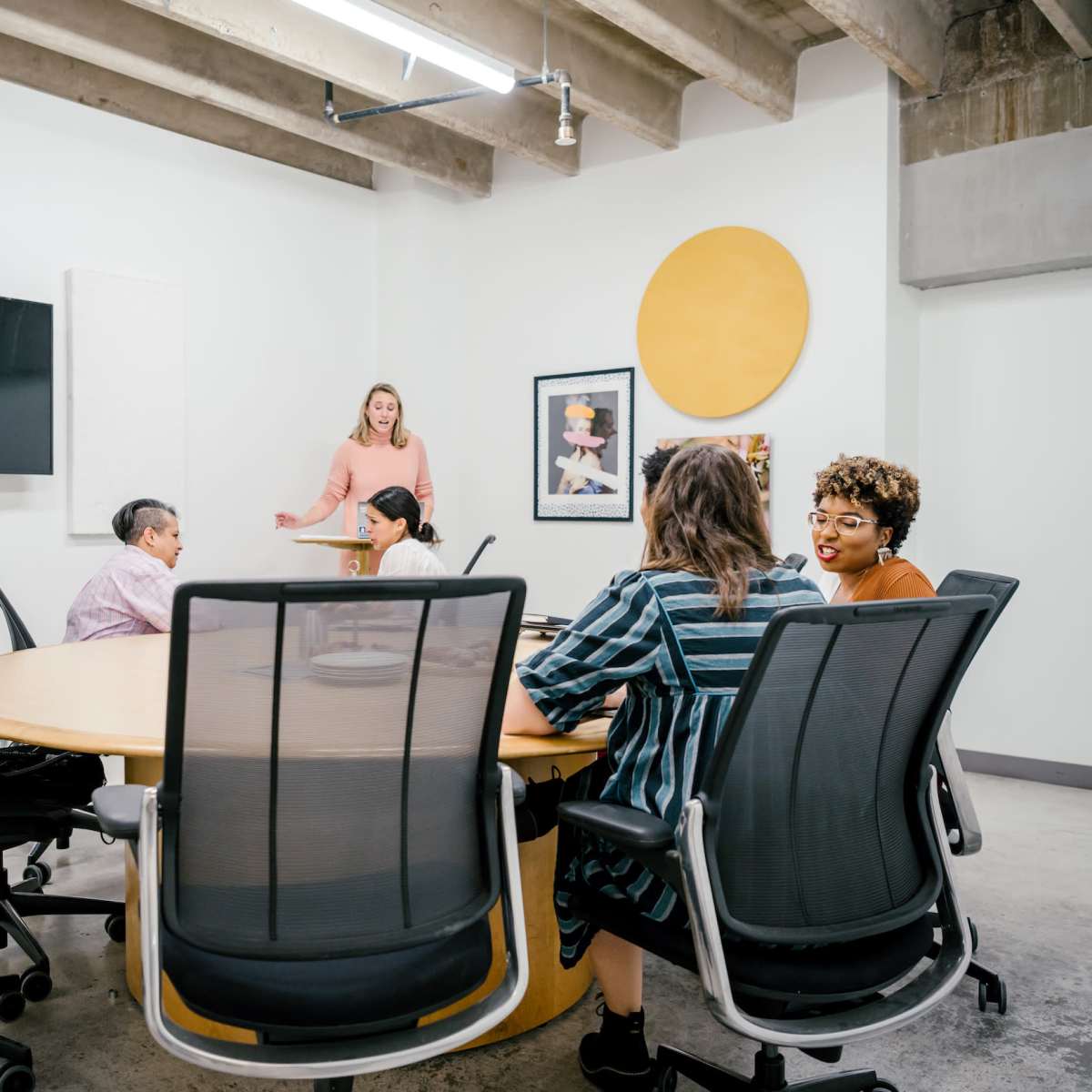A woman stands at the front of a conference room with a round table surrounded by several people engaged in discussion.