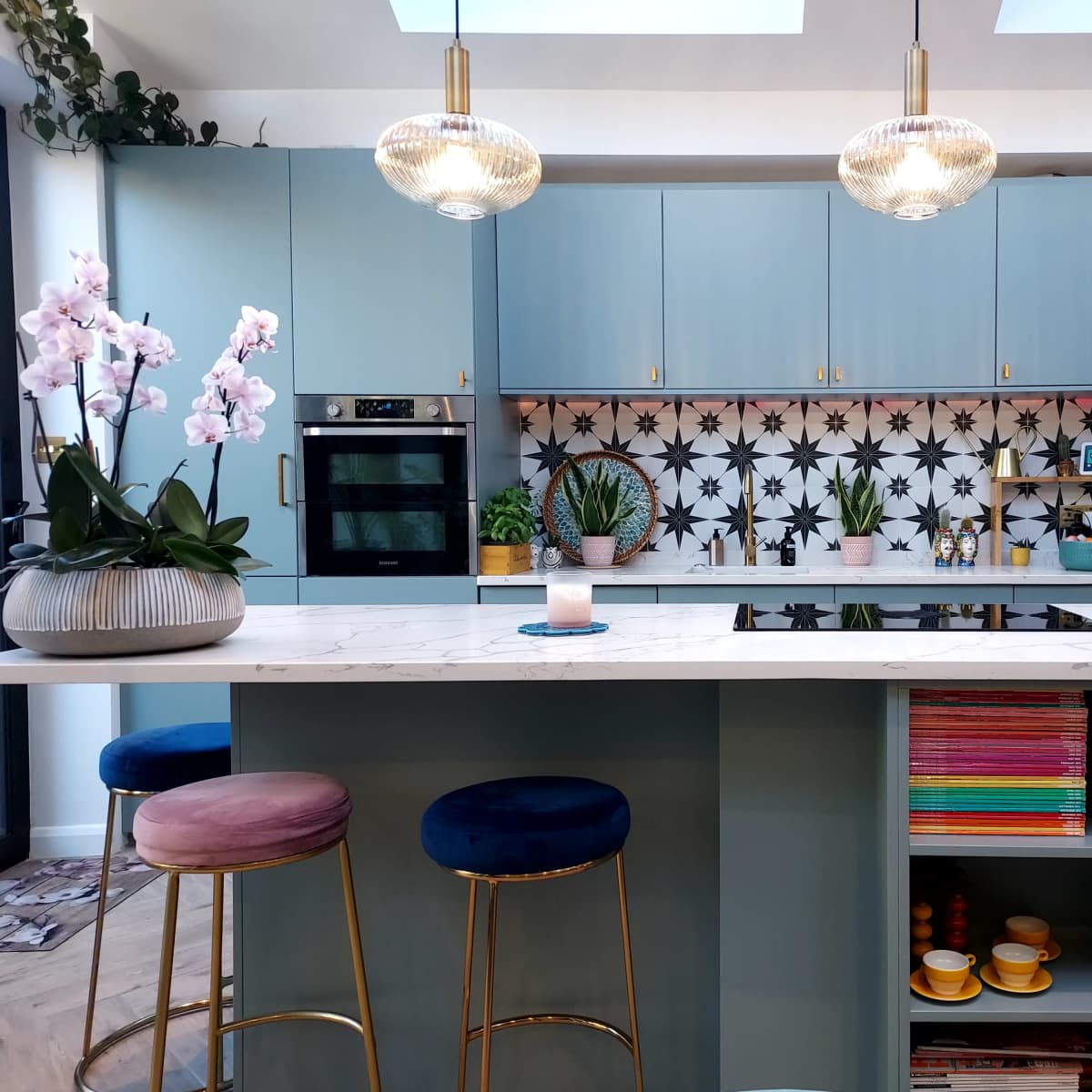 A modern kitchen features blue cabinets, a patterned backsplash, and a central island with bar stools and decorative plants.