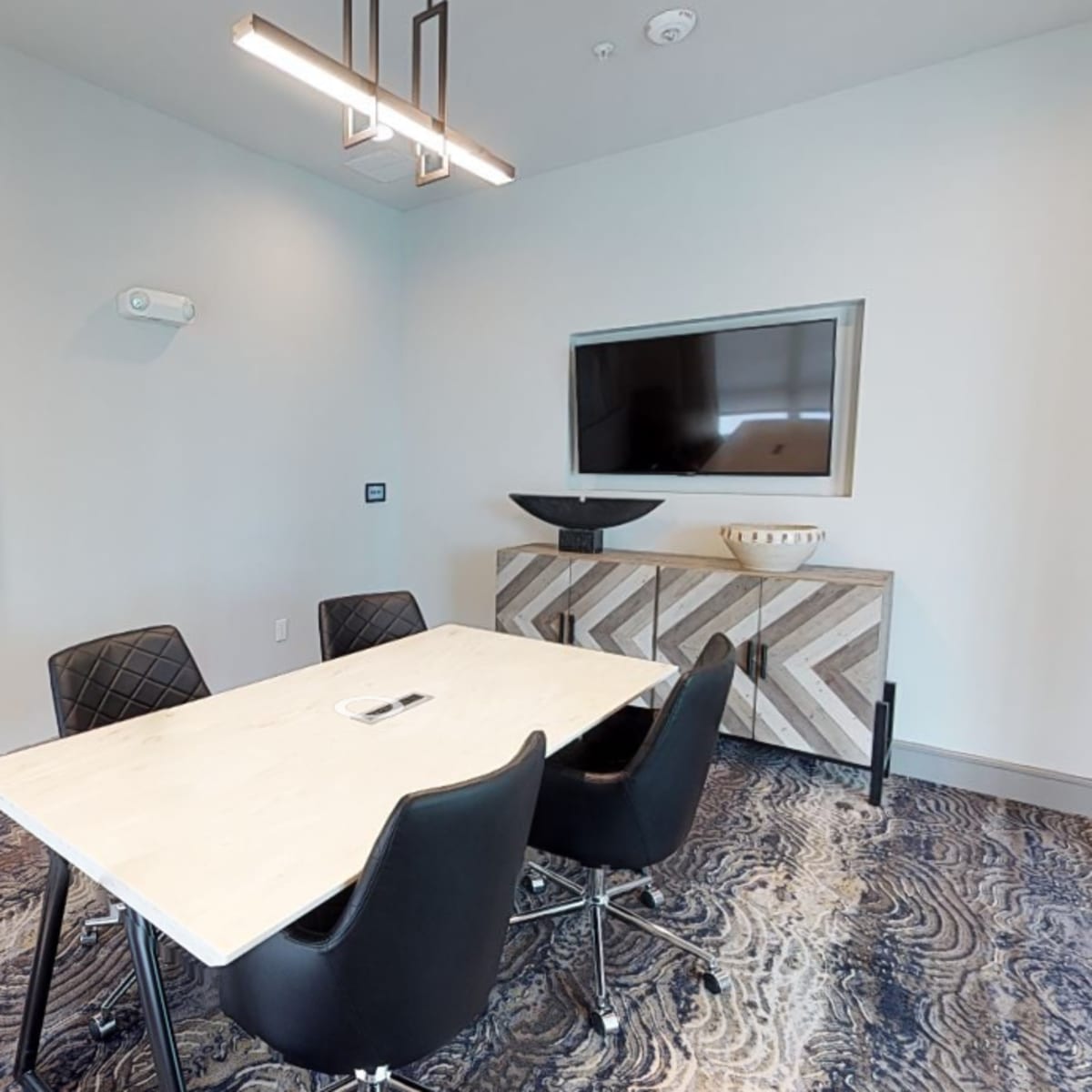 A modern conference room featuring a rectangular table surrounded by four black chairs, a wall-mounted TV, and abstract artwork.