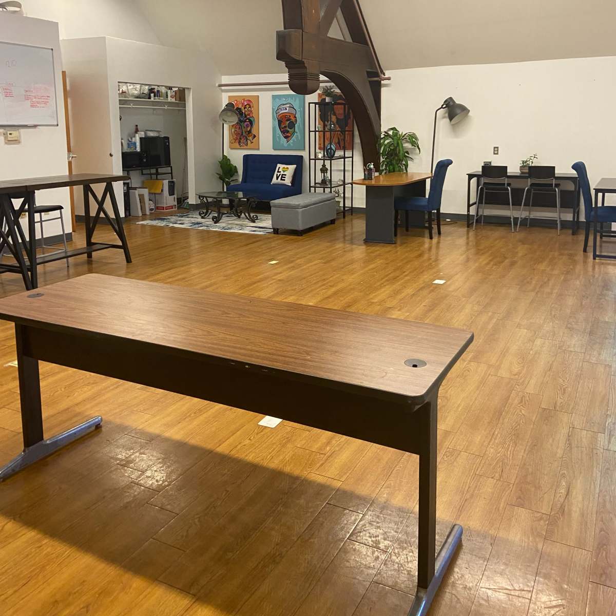 A spacious room with wooden flooring, featuring several tables and chairs, a sofa, and large beams on the ceiling.