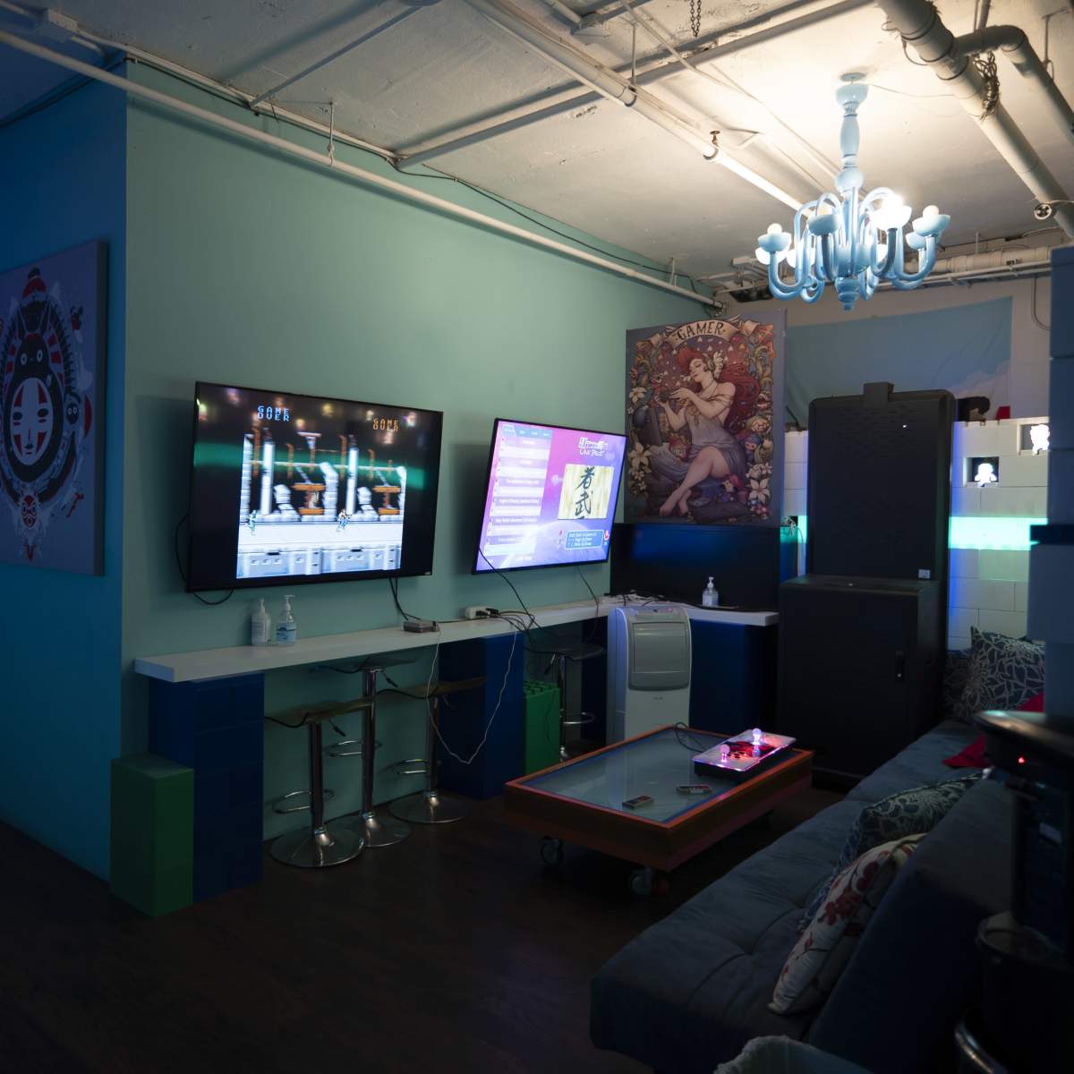 A dimly lit lounge area featuring two large televisions on the wall, a coffee table, and a cozy seating arrangement with a chandelier overhead.