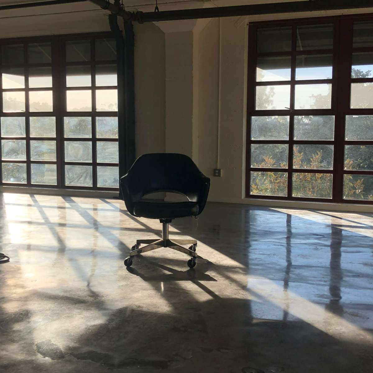 An empty office chair is positioned on a polished concrete floor, illuminated by sunlight streaming through large windows.