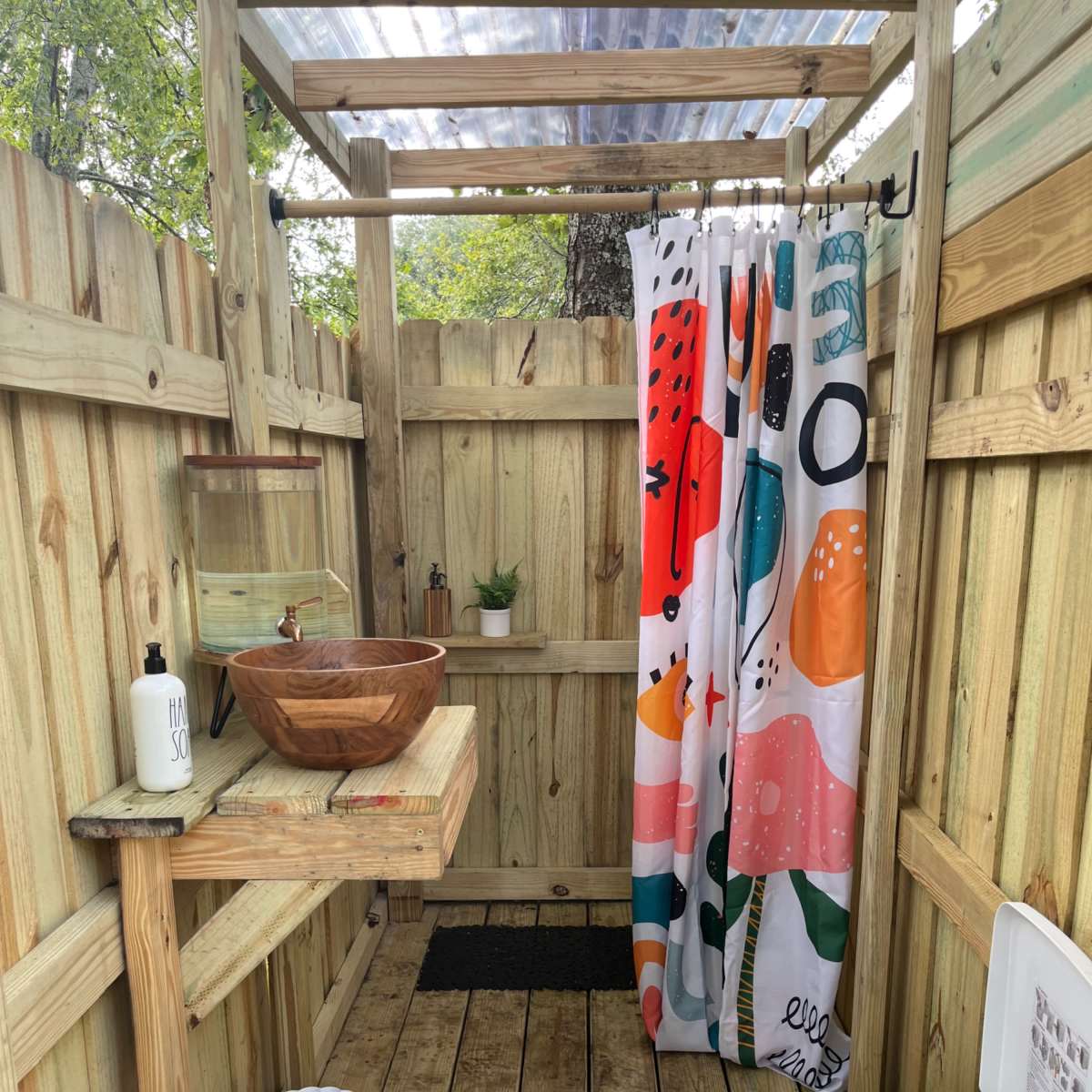 An outdoor shower area with wooden walls, a wooden sink with a bowl, a colorful shower curtain, and a portable toilet.