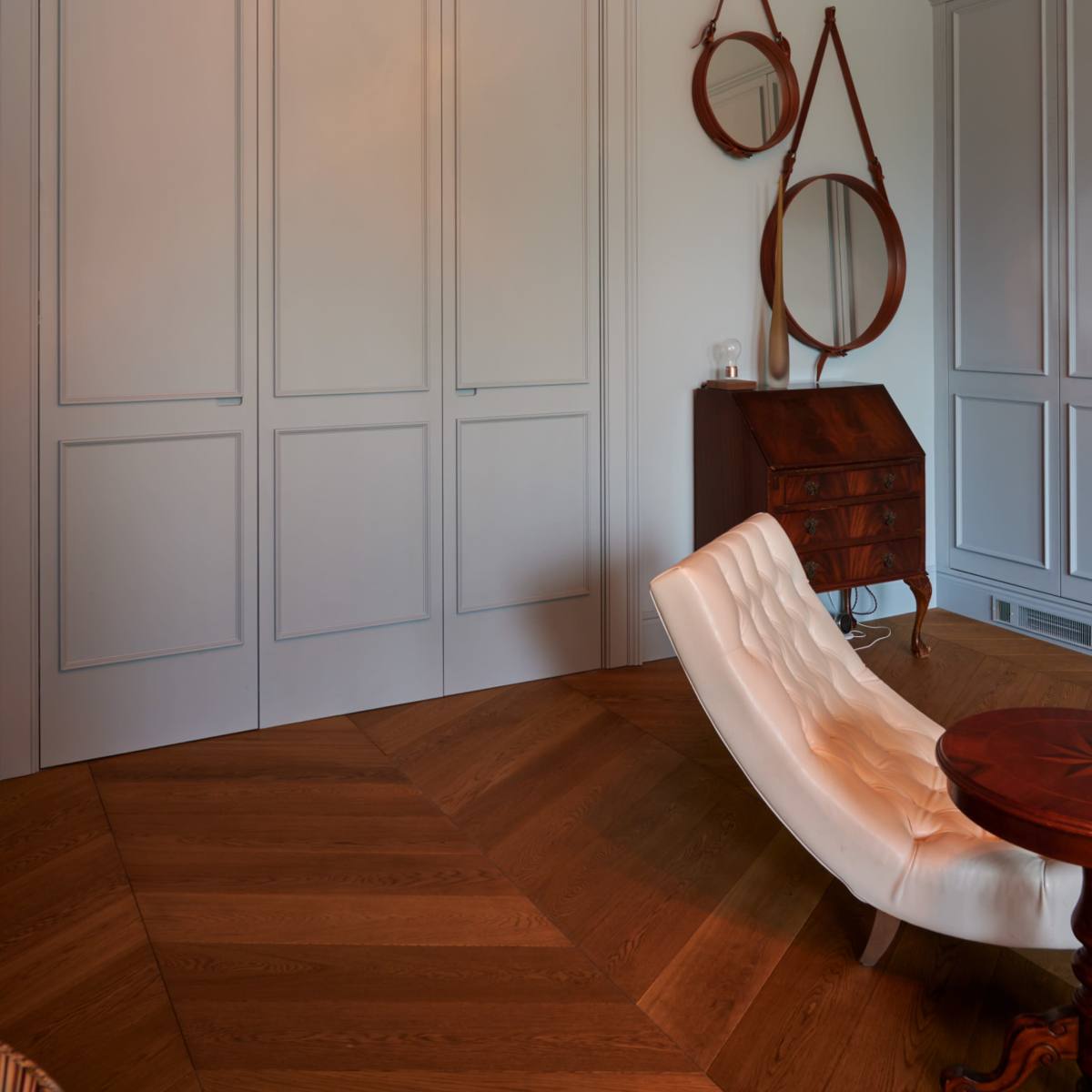 A room with light-colored walls, a wooden floor arranged in herringbone pattern, a small desk, and two round mirrors mounted on the wall.