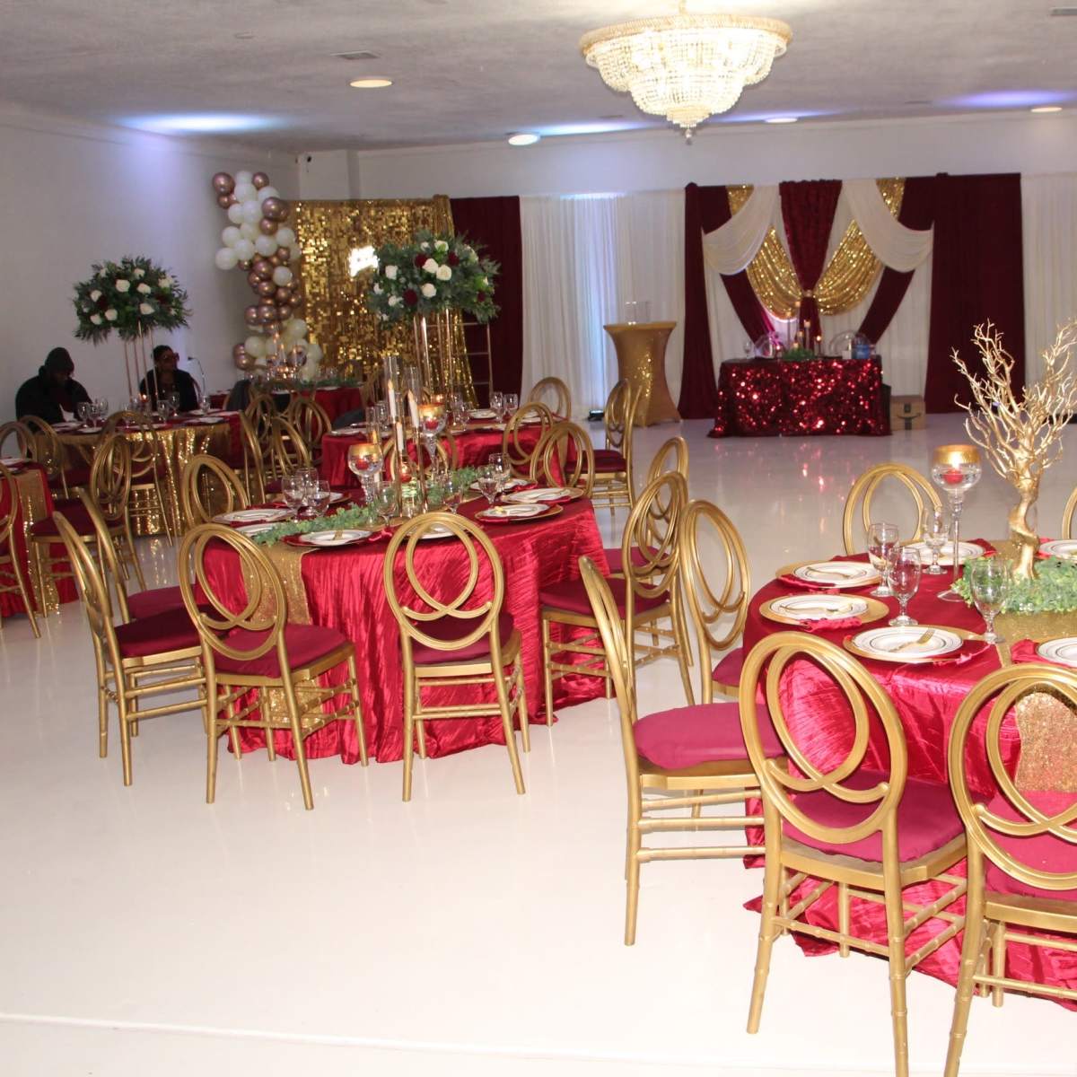 An elegantly decorated event space set for a formal gathering, featuring round tables with red tablecloths and gold chairs, adorned with floral centerpieces and glassware.