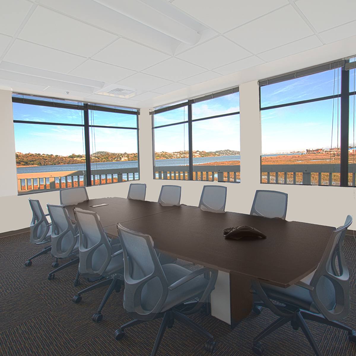 A conference room features a large table surrounded by chairs, with multiple windows offering a view of a landscape by the water.